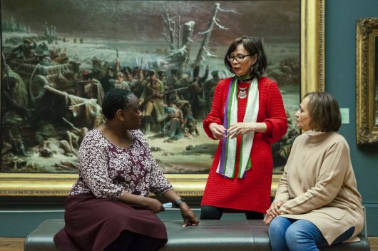 3 women in a gallery space, two are sat down on a bench and the woman in the middle is stood behind. In the background is a large framed painting