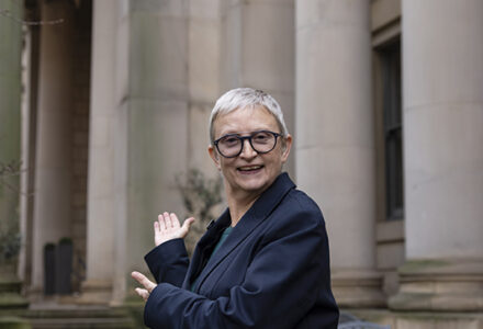 BSL tour guide Jennifer stood in the garden outside the gallery, she is looking at the camera and gesturing with her hands towards the entrance