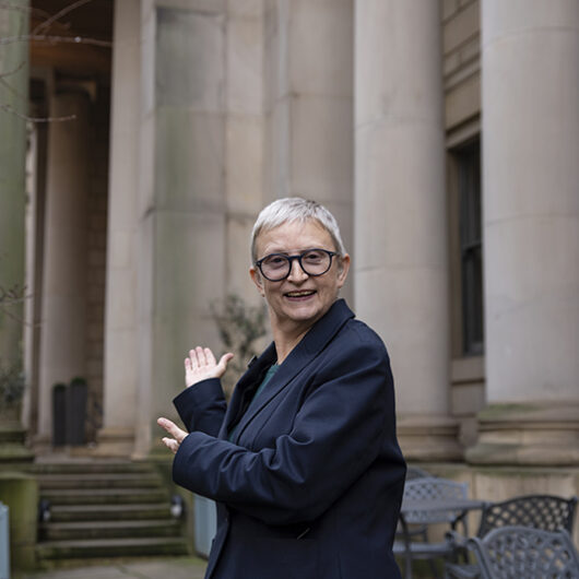 BSL tour guide Jennifer stood in the garden outside the gallery, she is looking at the camera and gesturing with her hands towards the entrance
