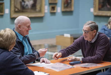 making conversation - a woman two older gentlemen sitting around a table, smiling whilst drawing in a gallery space