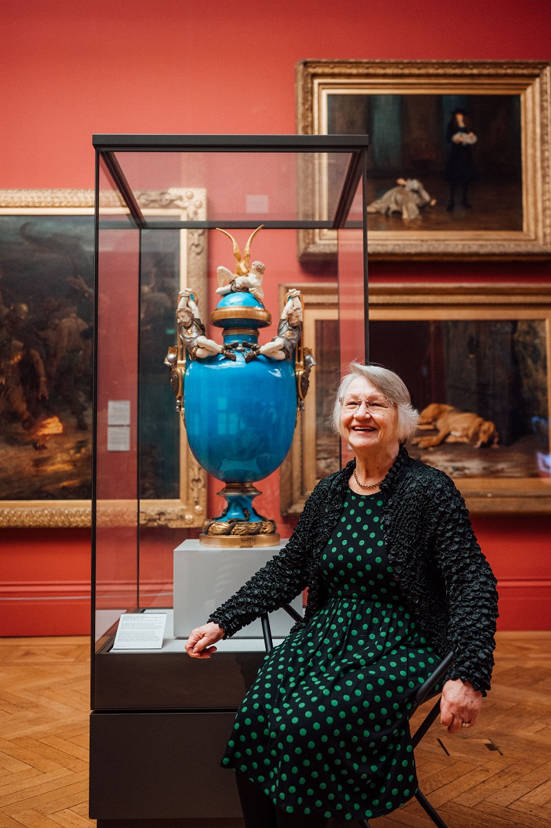 Portrait of Alina sat next to a large blue vase in a glass cabinet