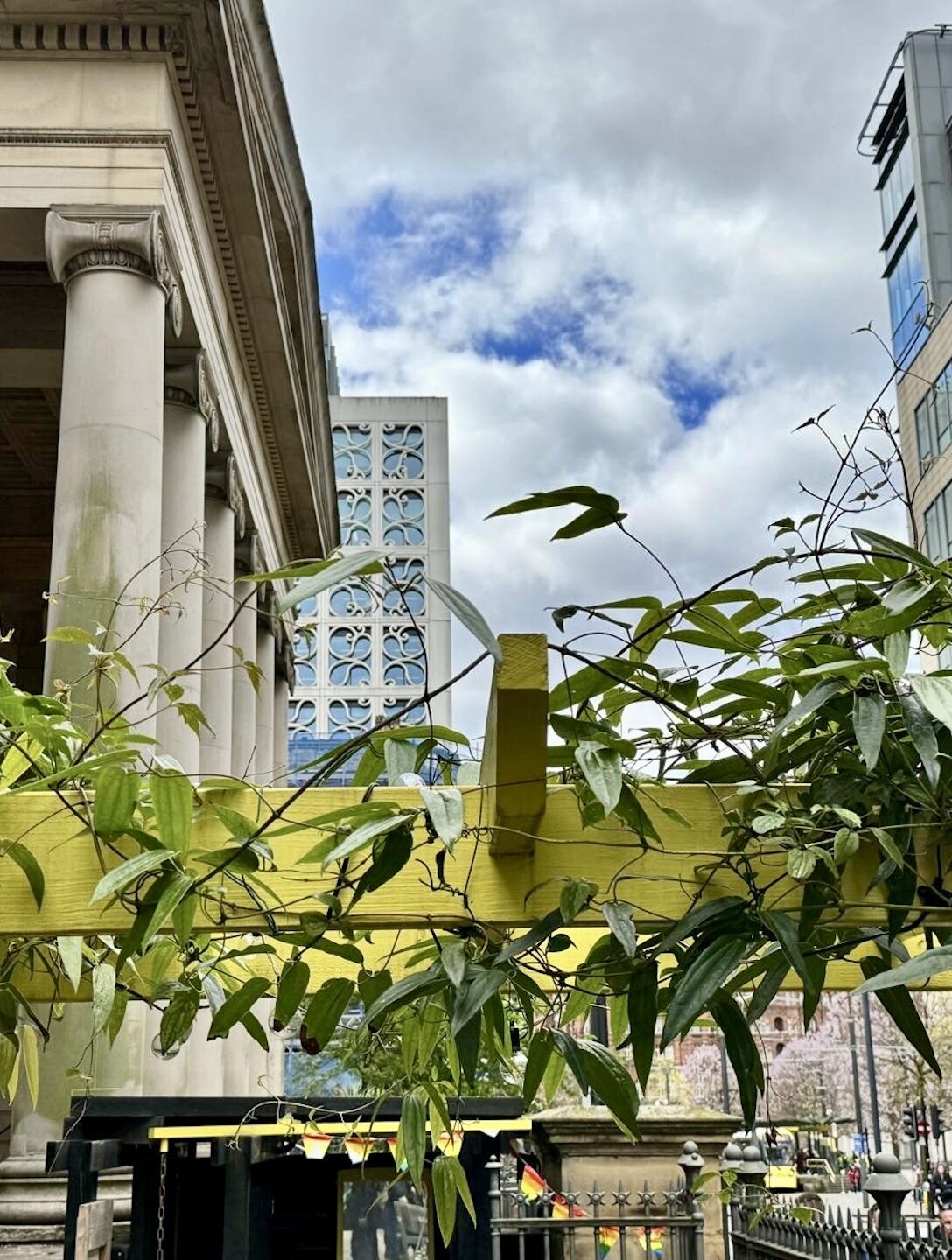 This image contrasts the old and new architectural styles in a city setting. In the foreground, there is an ornate, historical stone building with decorative columns and arches, representing traditional architecture. However, directly behind it stands a modern, glass and steel high-rise building with a geometric design, showcasing contemporary architectural styles. The juxtaposition is further highlighted by lush, green foliage in the foreground, adding a natural element amidst the urban landscape. The cloudy sky and presence of pedestrians contribute to the dynamic, city atmosphere captured in the scene.