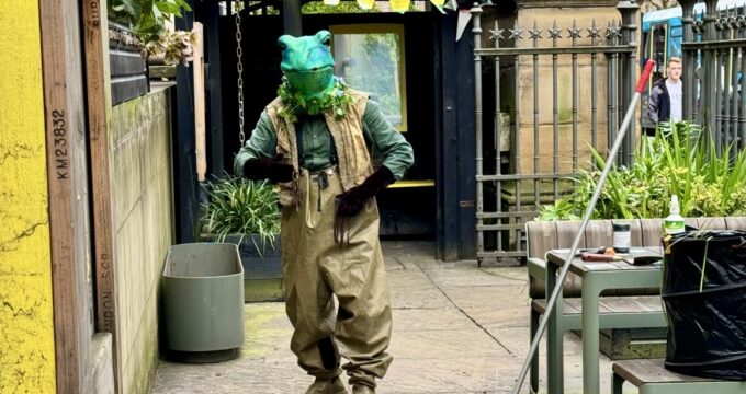 a man in a green frog mask is walking and lceaning the sidewalk