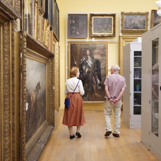 A man and woman walking away from the camera, looking at a gallery space, the walls are filled with framed paintings