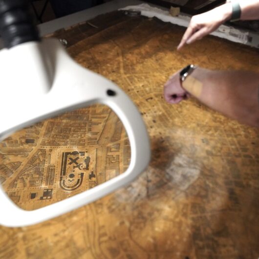 detail of an antique map of Manchester, one section enlarged through a magnifying glass. Two hand point to a section of the map to the right