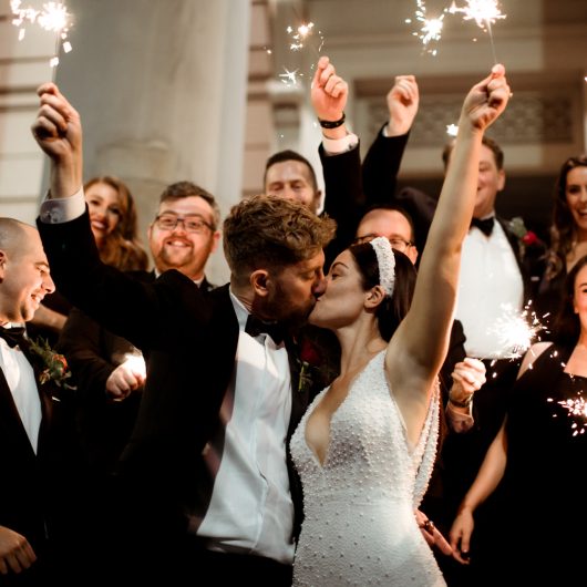 Married couple on the steps of the gallery kissing. Guests are holding sparklers in the air