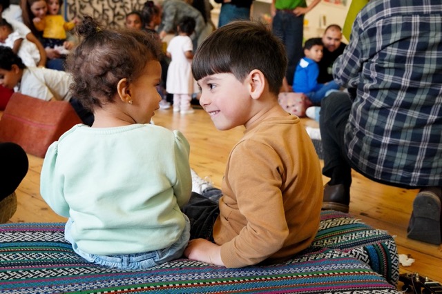 Two small children sat on a cushion together, looking at each other, in the background are other families attending the workshop