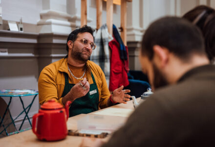 A man in his 30s wearing round glasses and a yellow sweatshirt talking to a couple on the other side of the table, possibly about the teapots sitting in front of them