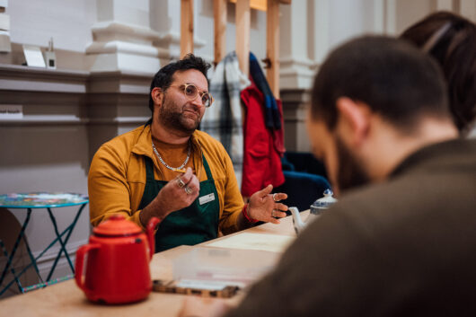 A man in his 30s wearing round glasses and a yellow sweatshirt talking to a couple on the other side of the table, possibly about the teapots sitting in front of them