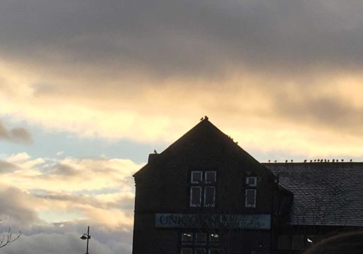 A photograph of a sunset. The background is grey orange, yellow, light and dark grey. There is a silhouette of a building with a part sloping and part flat roof towards the bottom right corner.
