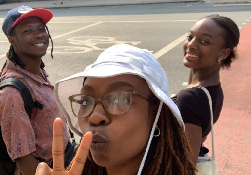 The image show the collective of young Black creatives in Manchester standing in front of a road, smiling at the camera and finger gesturing the number three.