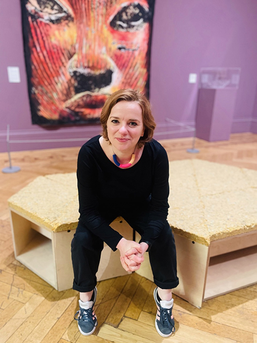 Portrait of Louise Thompson - a woman with short ginger hair sat on a bench leaning forwards with her hands clasped together and looking at the camera. Sat in a gallery space with purple walls and a large painting hanging behind.