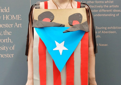 Bust of a person wearing a paper dress. Consisting of a Cuban flag with two black eyes and red pupils and shoulder pads with brown fringes.