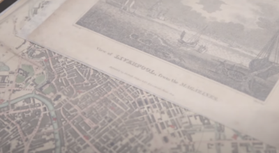 The image shows a close-up view of old documents or maps. In the foreground, there's a detailed city map with streets and buildings visible. The map appears to be of an older style, possibly historical. Above the map, there's what looks like a panoramic sketch or engraving of a cityscape. The documents have a faded, sepia-toned appearance, suggesting they are quite old. The overall composition gives the impression of looking at historical documents or archival materials related to urban planning or city history.