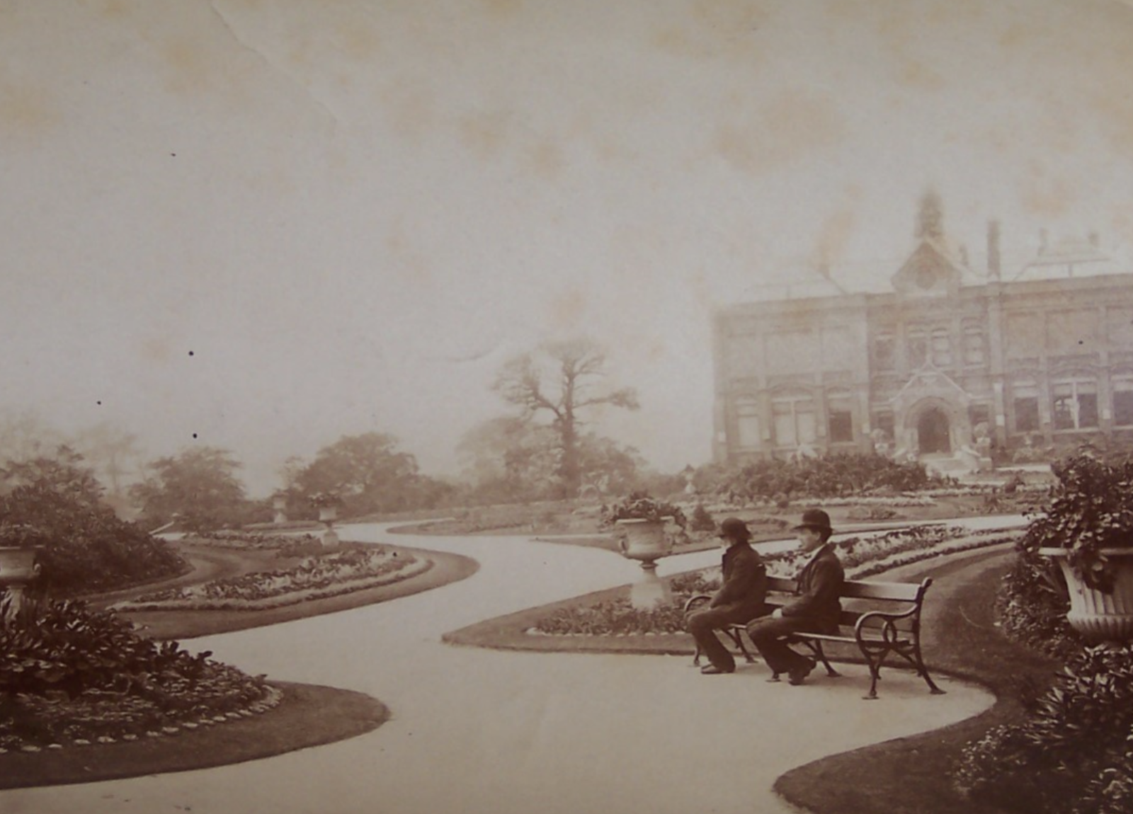 A black and white photograph of a Victorian park, in the centre, two people sit on a bench at the edge of a winding path that leads to Queens Park gallery in the background
